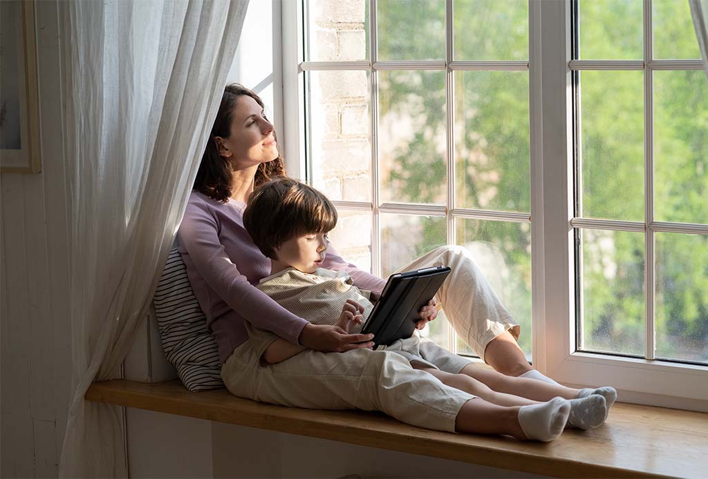 Caring mom engaging with small preschool son on weekend relaxed sitting on windowsill hugging kid playing on digital tablet and enjoy day sunlight from outside. Parent and child at home on quarantine