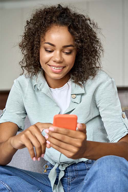 Happy young African American woman looking at cell phone relaxing on sofa at home, using online mobile apps tech, doing mobile shopping buying on mobile phone, checking social media, texting.